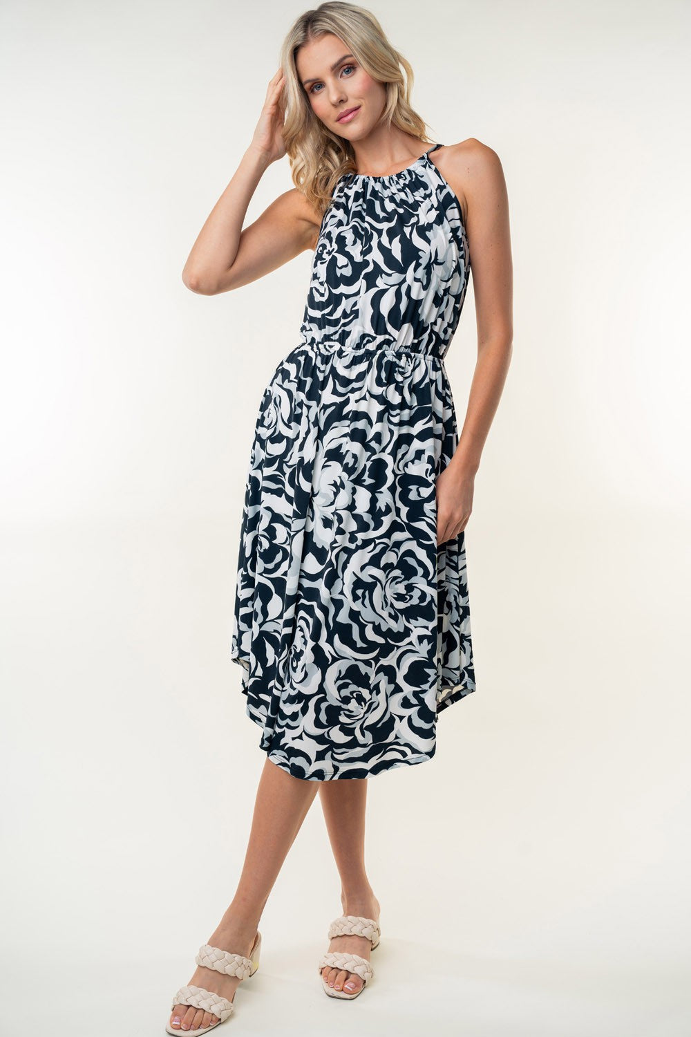 The person wearing a White Birch Tied Ruched Floral Sleeveless Knee Length Dress, featuring a black and white floral pattern, stands and smiles against a neutral background.