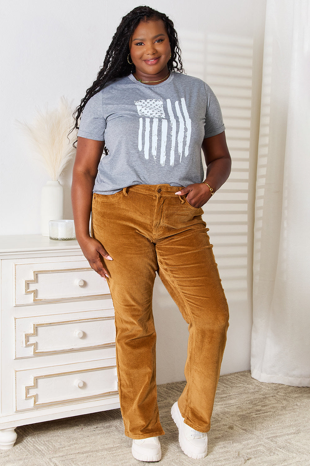 A woman with long dark hair is wearing a Simply Love US Flag Graphic Cuffed Sleeve T-Shirt and brown pants as she stands indoors against a light-colored background.