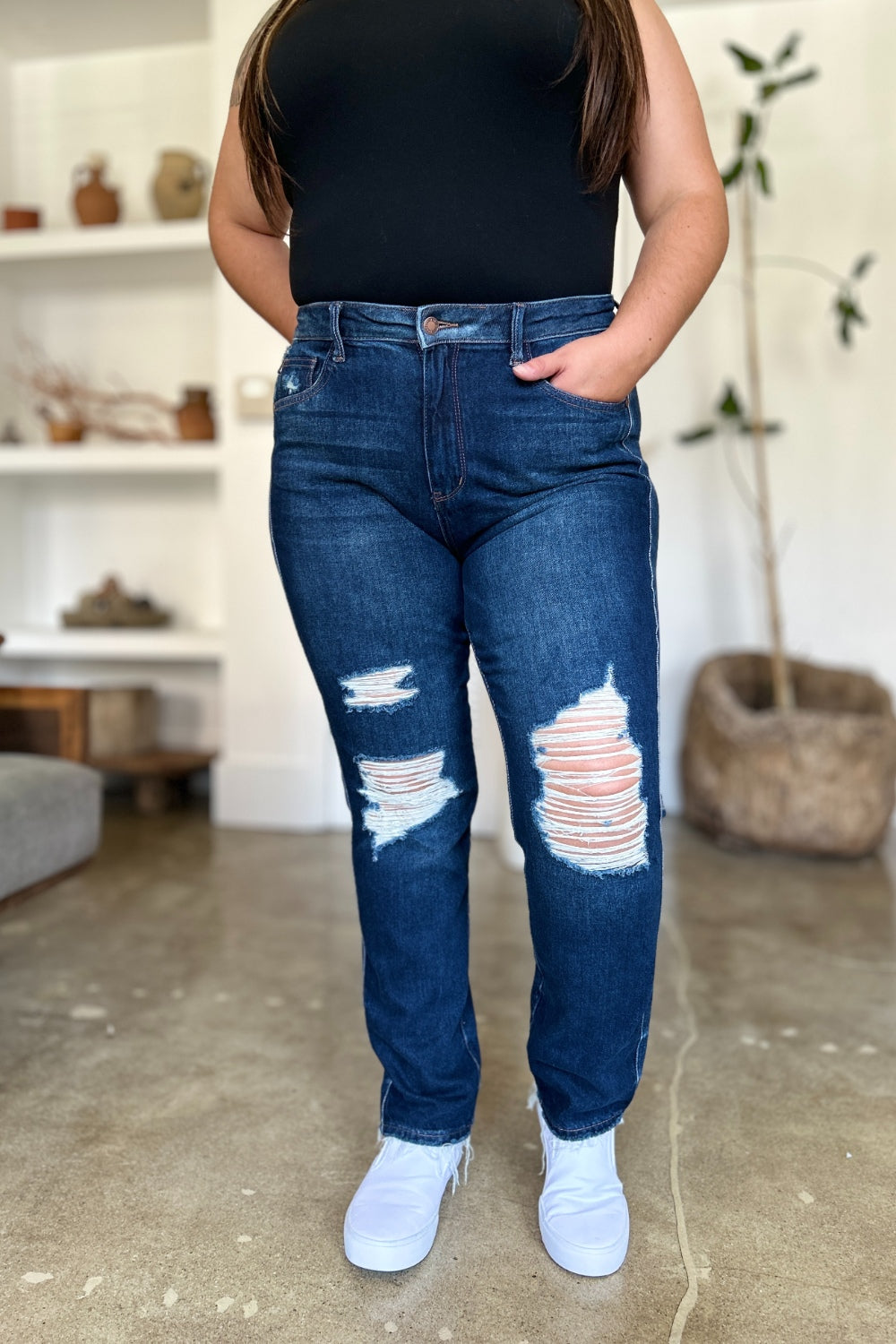 A woman in a black sleeveless top and Judy Blue Full Size High Waist Rigid Magic Heavy Destroy Straight Jeans stands indoors with her hands in her pockets. The background features a plant and shelves adorned with decor items.