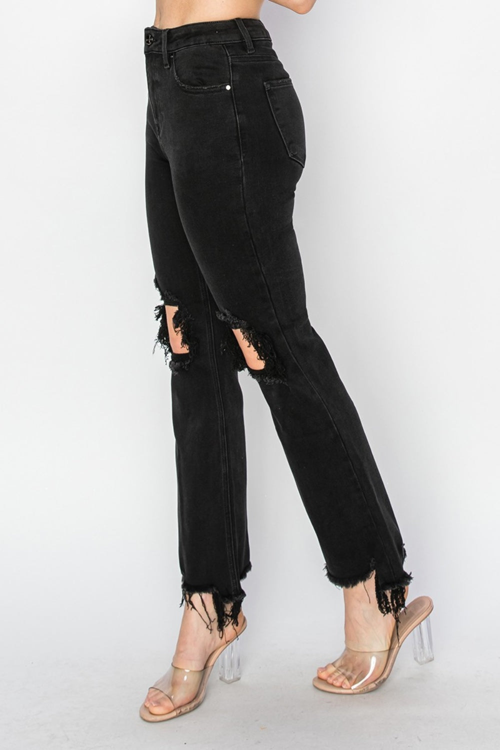 Rear view of a person wearing RISEN Full Size Distressed Raw Hem Jeans with Pockets in black, paired with clear-heeled sandals against a plain background.