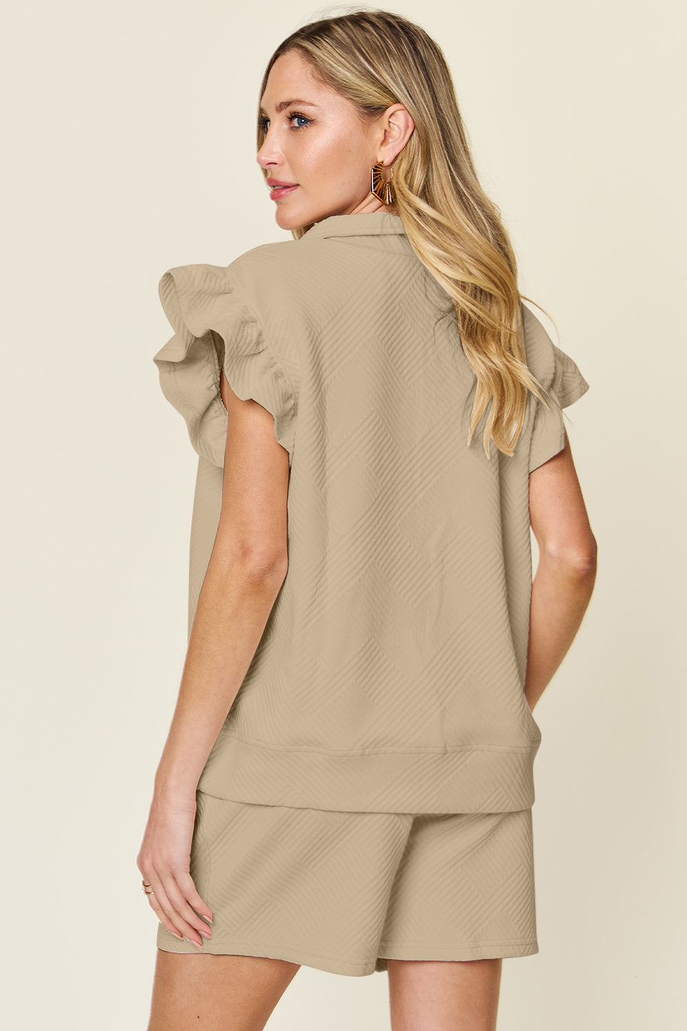 Against a plain background, a woman with blonde hair poses confidently with one hand in her pocket, highlighting the chic style of her Double Take Full Size Texture Flounce Sleeve Top and Drawstring Shorts Set.