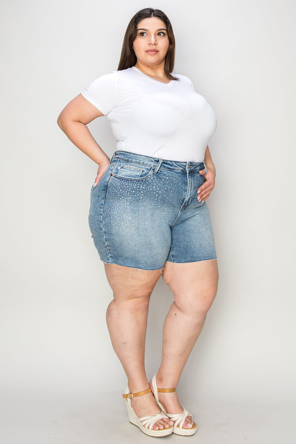 A woman wearing a white T-shirt, Judy Blue Full Size High Waist Rhinestone Decor Denim Shorts, and white sandals poses against a plain background.