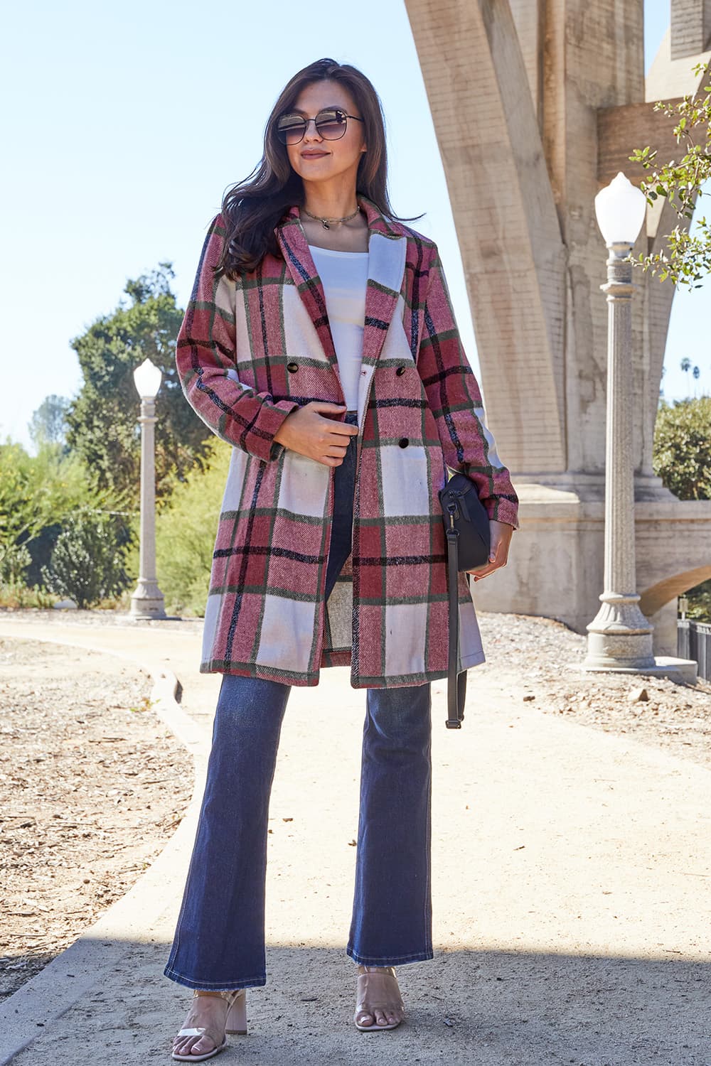 A woman stands on a sidewalk wearing the Double Take Full Size Plaid Button Up Lapel Collar Coat in pink and white over a white top and blue jeans, with her right hand lightly touching her hair. The polyester fabric of the coat makes it durable and perfect for an easy machine wash cold.