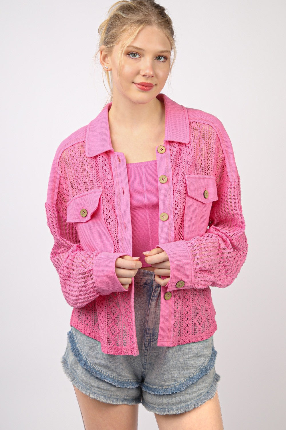 A person wearing a bright pink, long sleeve button-up lace shirt with drop shoulders and high-waisted denim shorts stands against a plain background.