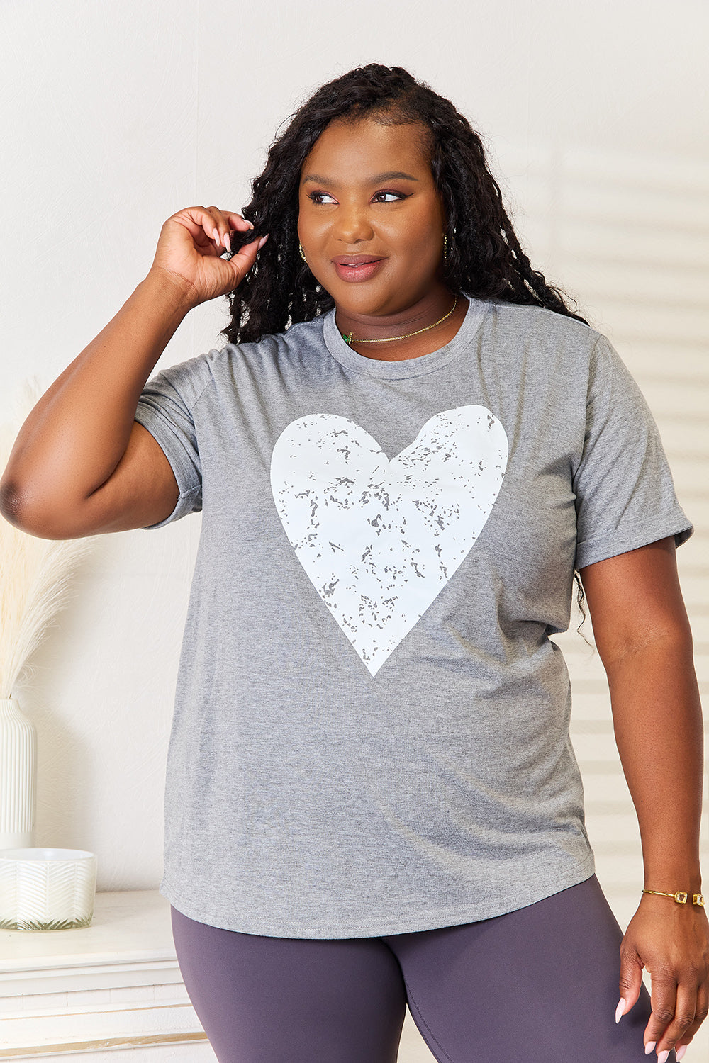 A woman with long hair stands indoors, smiling and touching her hair, wearing the trendy Simply Love Heart Graphic Cuffed Short Sleeve T-Shirt in gray.