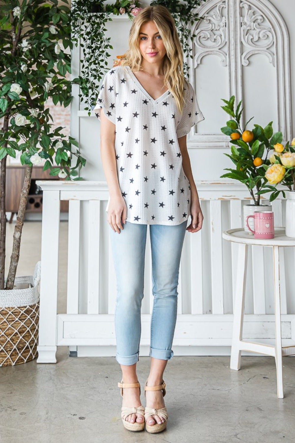 A person with long blonde hair is wearing the Heimish Full Size Star Print Short Sleeve V-Neck Waffle Knit T-Shirt. They are standing indoors near a plant.