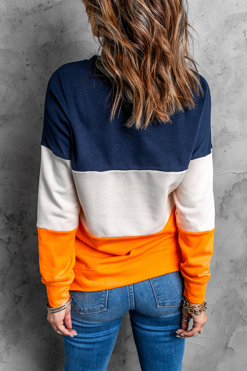 Person with wavy, shoulder-length hair wearing a Colorblock Orange Contrast Stitching Sweatshirt with Slits, paired with blue skinny fit jeans, standing with back to the camera against a gray textured wall.