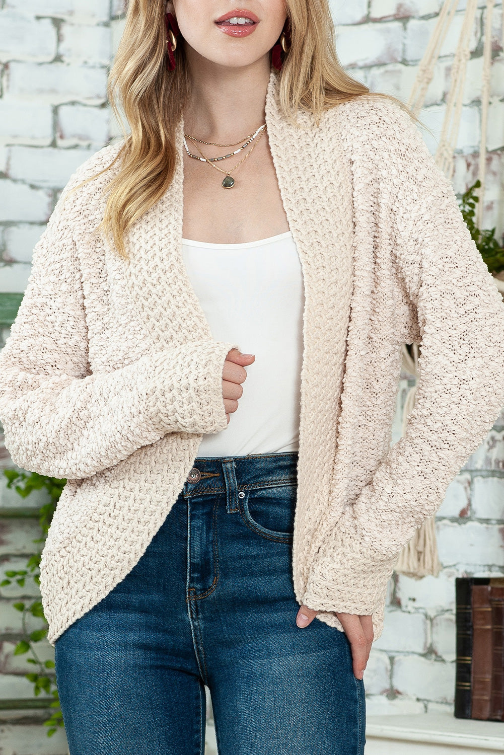 A woman in an Apricot Popcorn Knit Open Front Cardigan and jeans stands against a brick wall, looking to the side with a slight smile.