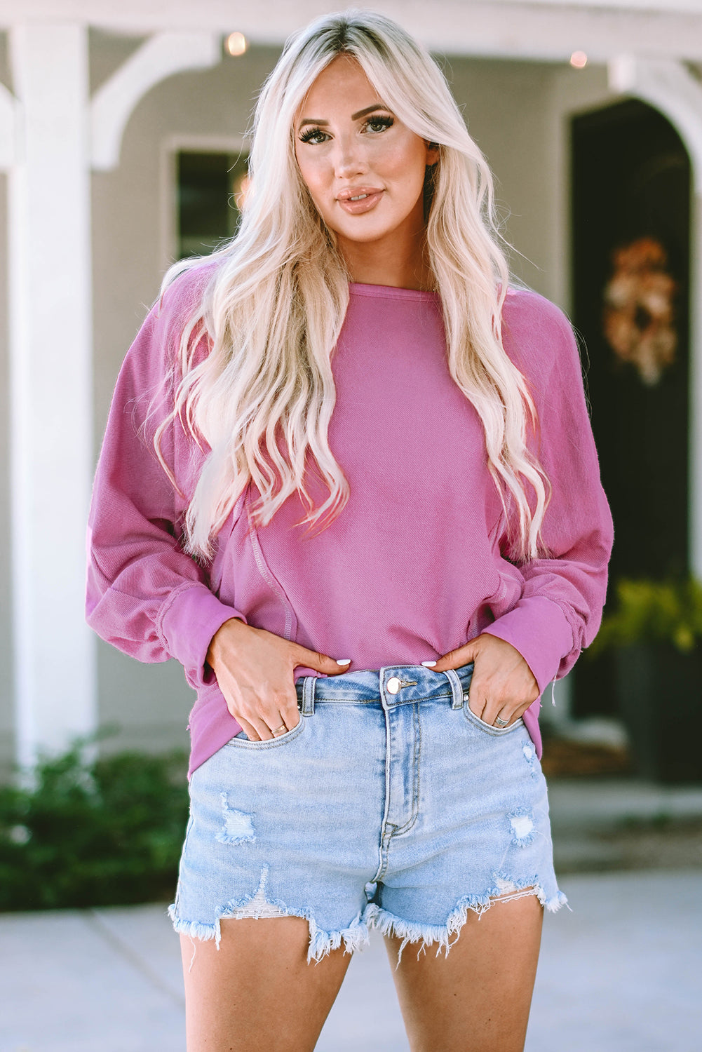 A person with long blonde hair is seen from the back wearing a Pink Exposed Seam Round Neck Terry Pullover and distressed denim shorts. The trendy look complements the casual outfit as they stand outdoors in front of a building with a window and a lantern.