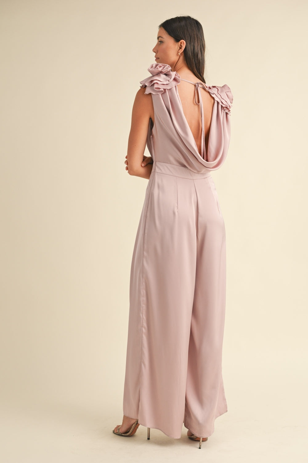 A woman stands against a plain background wearing the MABLE 3D Floral Applique Deep Cowl Neck Jumpsuit, which features an open back and wide-leg pants, paired with open-toed heels.