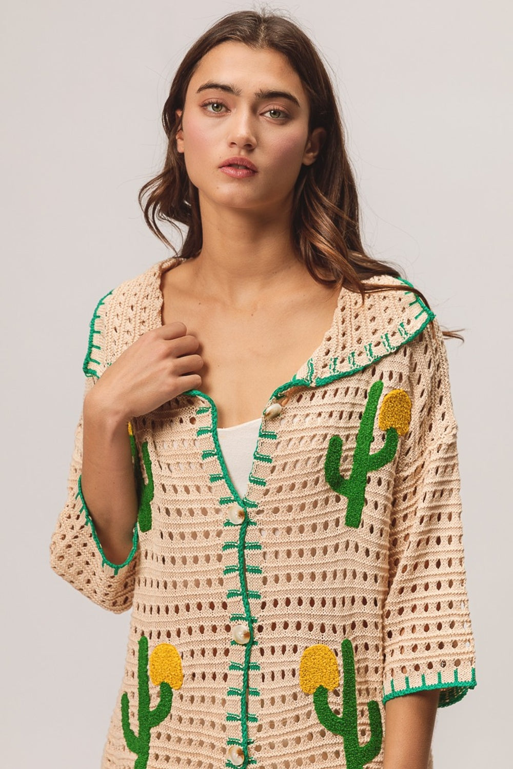 A woman wearing the BiBi Edge Stitched Cactus Patch Sweater Cardigan, featuring beige fabric with green cactus designs, layered over a white top and paired with white frayed shorts, stands against a plain background.