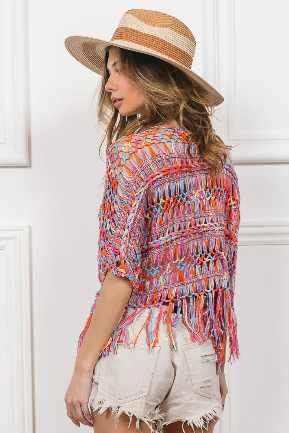 A woman wearing a BiBi Open Front Fringed Crop Knit Cardigan, a white bandeau, distressed white shorts, and a wide-brimmed straw hat stands against a white paneled background, smiling with one hand on her hat.