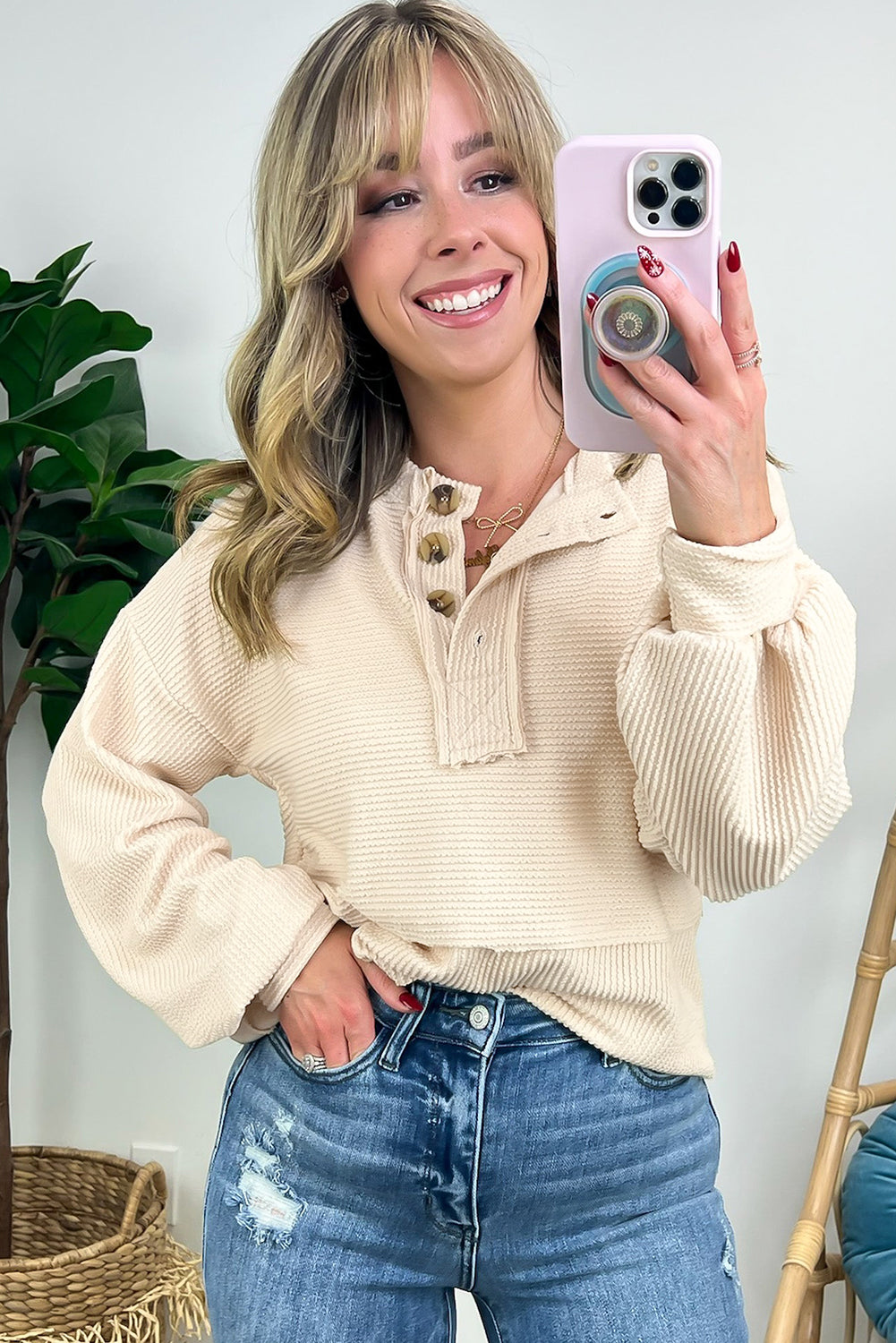 Person wearing the Apricot Corded Texture Lantern Sleeve Buttons Henley Sweatsuit and blue jeans, taking a mirror selfie with a smartphone. Hair is straight and shoulder-length; they stand in front of a white wall and a wooden chair.