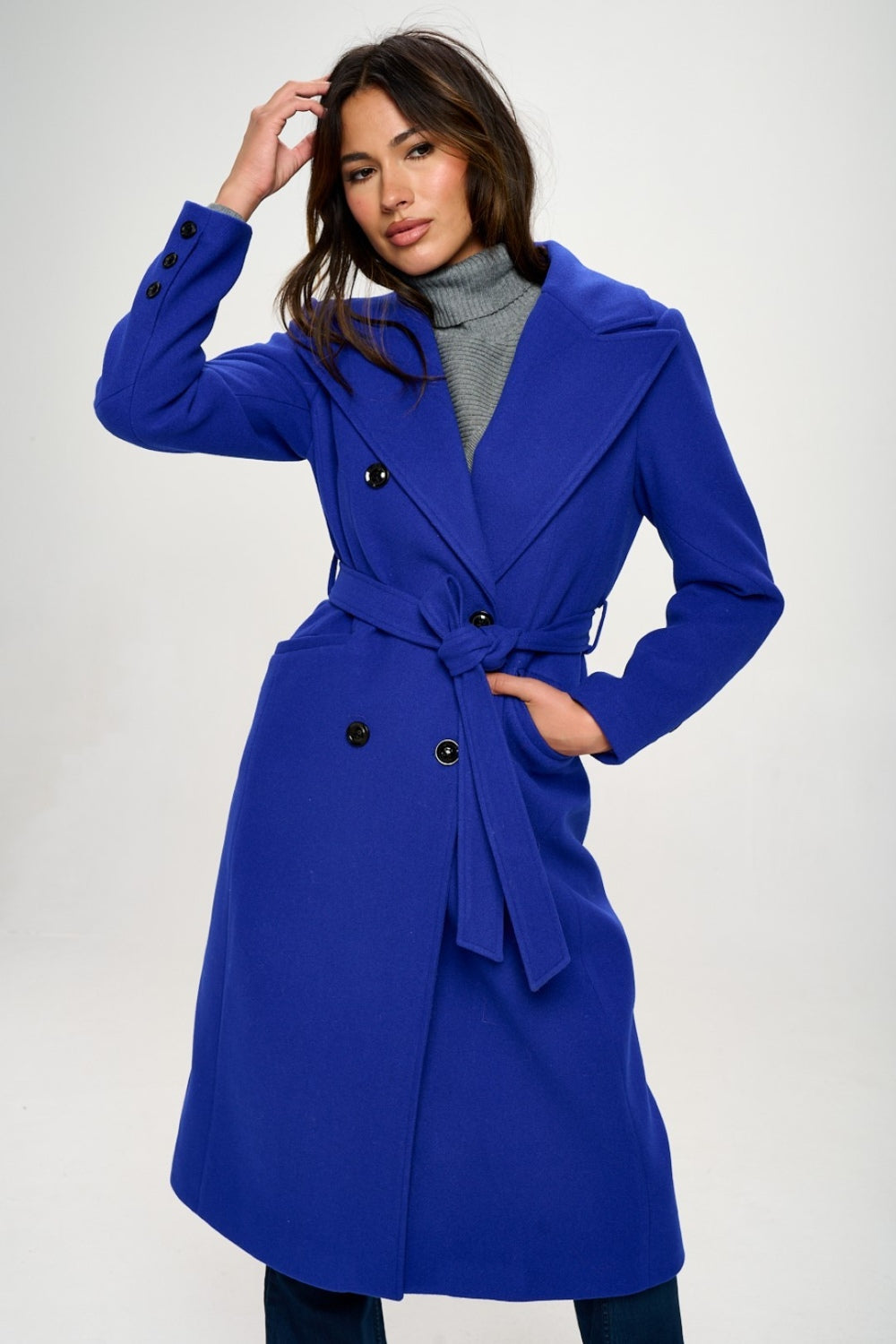 Person wearing the Coalition LA Double-Breasted Longline Coat with Belt, made from vegan wool in blue, over a grey turtleneck, posing against a plain background.