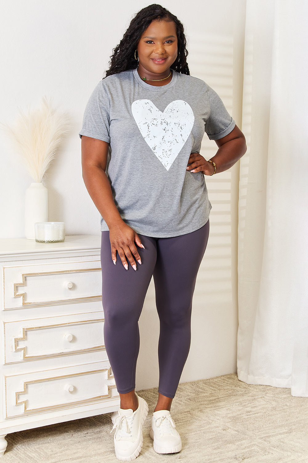 A woman with long hair stands indoors, smiling and touching her hair, wearing the trendy Simply Love Heart Graphic Cuffed Short Sleeve T-Shirt in gray.