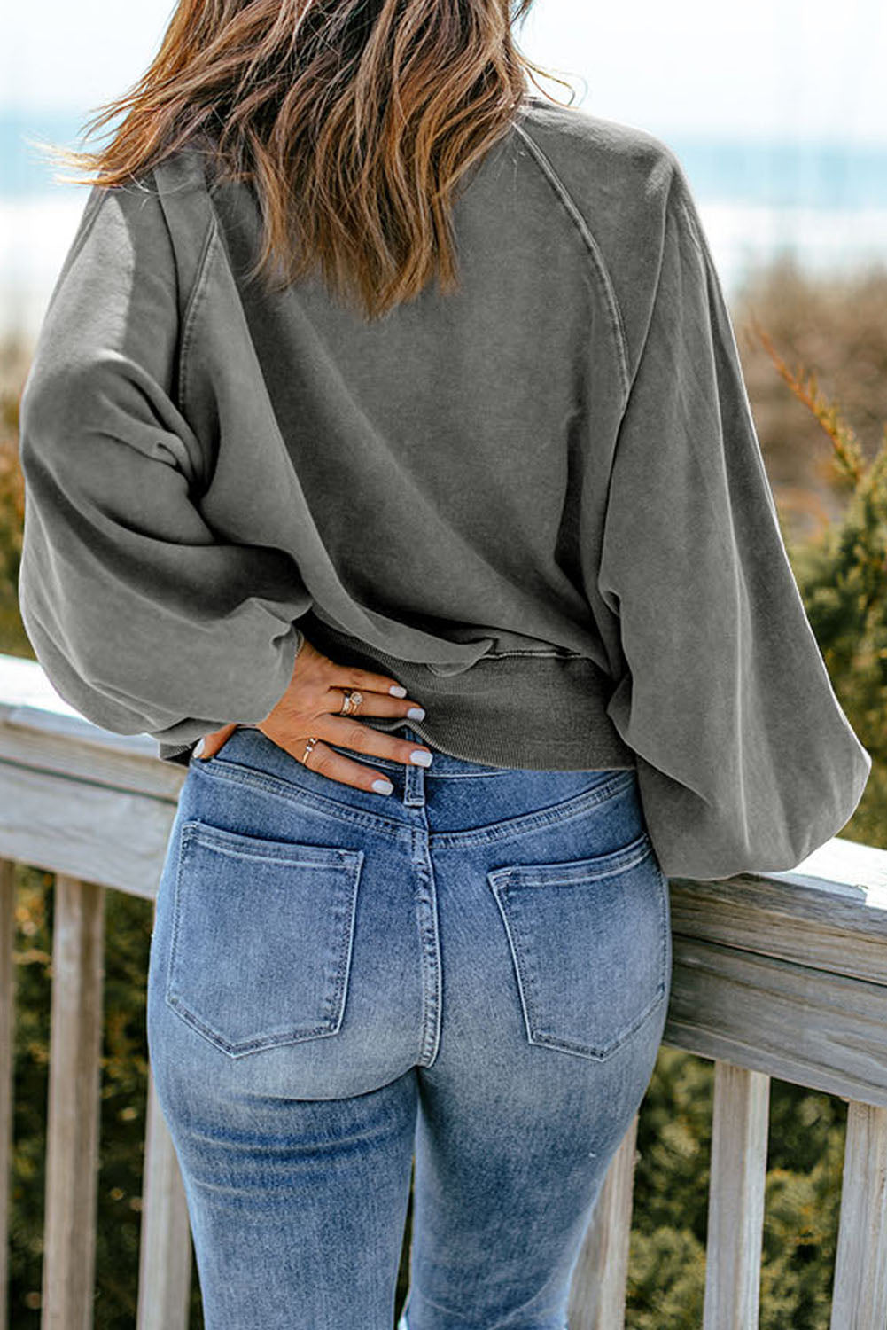 A person with long hair stands outdoors, facing away, wearing a chic **Gray Washed Snap Buttons Lantern Sleeve Pullover Sweatshirt** and blue jeans, resting their hands on a wooden railing.