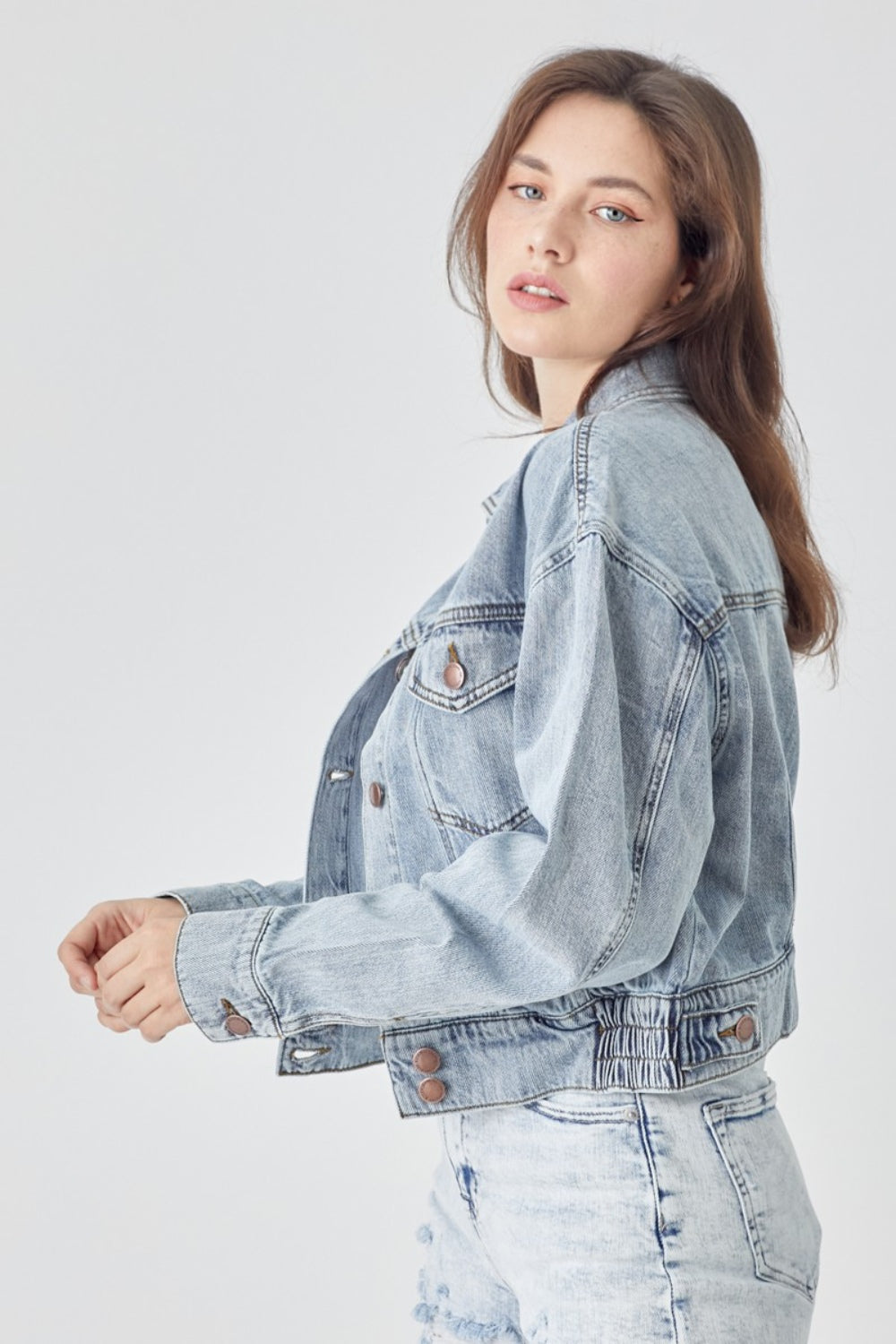 A woman wearing the RISEN Full Size Button Down Cropped Denim Jacket over a white top and distressed denim shorts stands against a plain background, showcasing versatile outerwear perfect for any casual occasion.