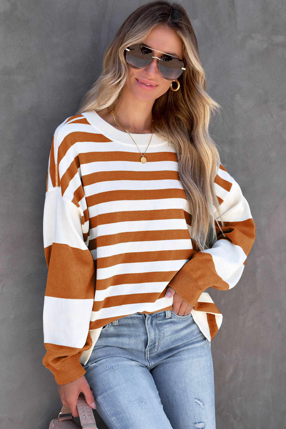 A person wearing sunglasses, gold earrings, a gold necklace, a Brown Stripe Drop Shoulder Striped Pullover Sweatshirt in white and orange stripes, and light blue jeans stands against a gray backdrop.