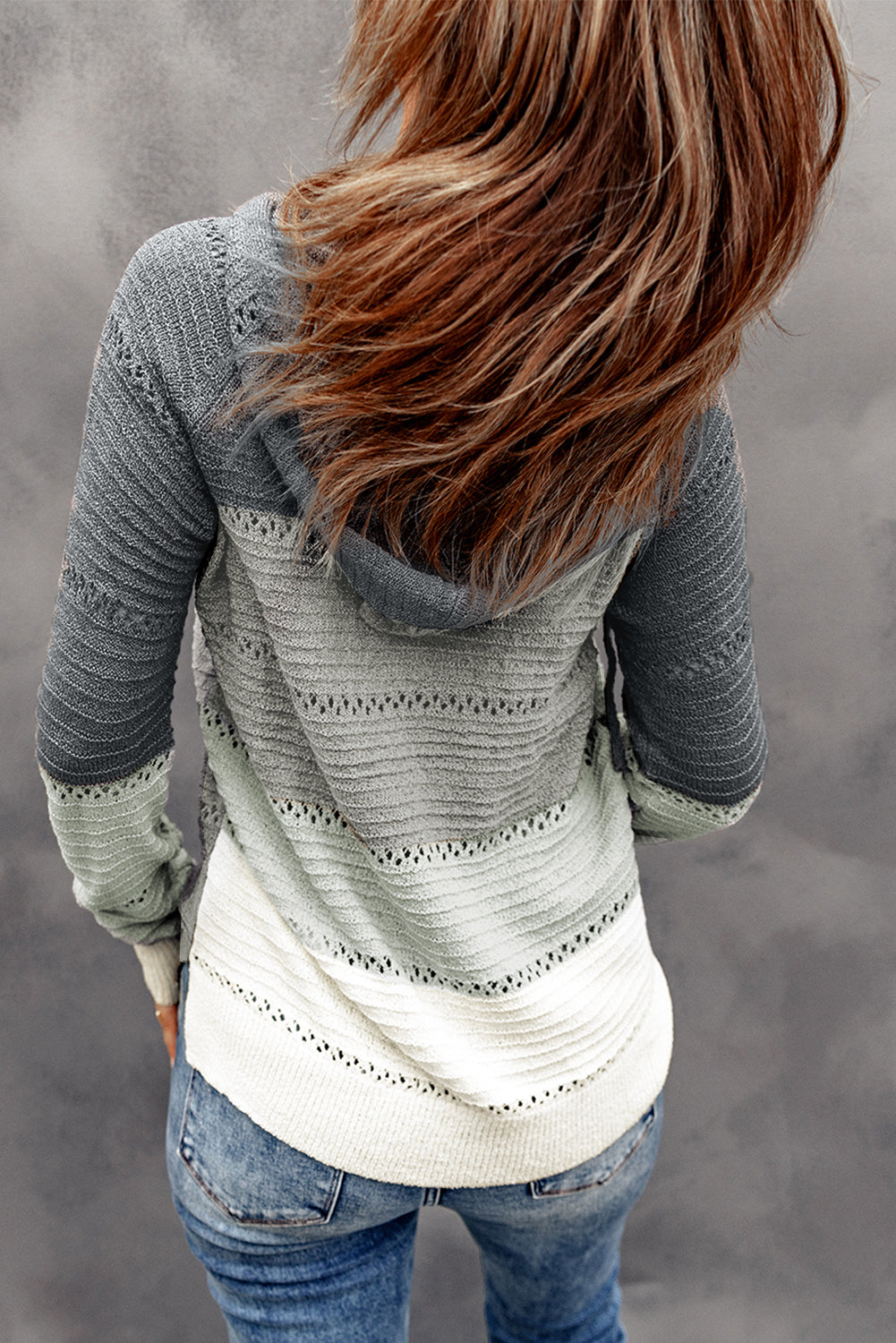 Rear view of a person with long hair wearing the Beach Bonfire Knitted Hoodie and blue jeans, showcasing one of our casual styles against a gray background.