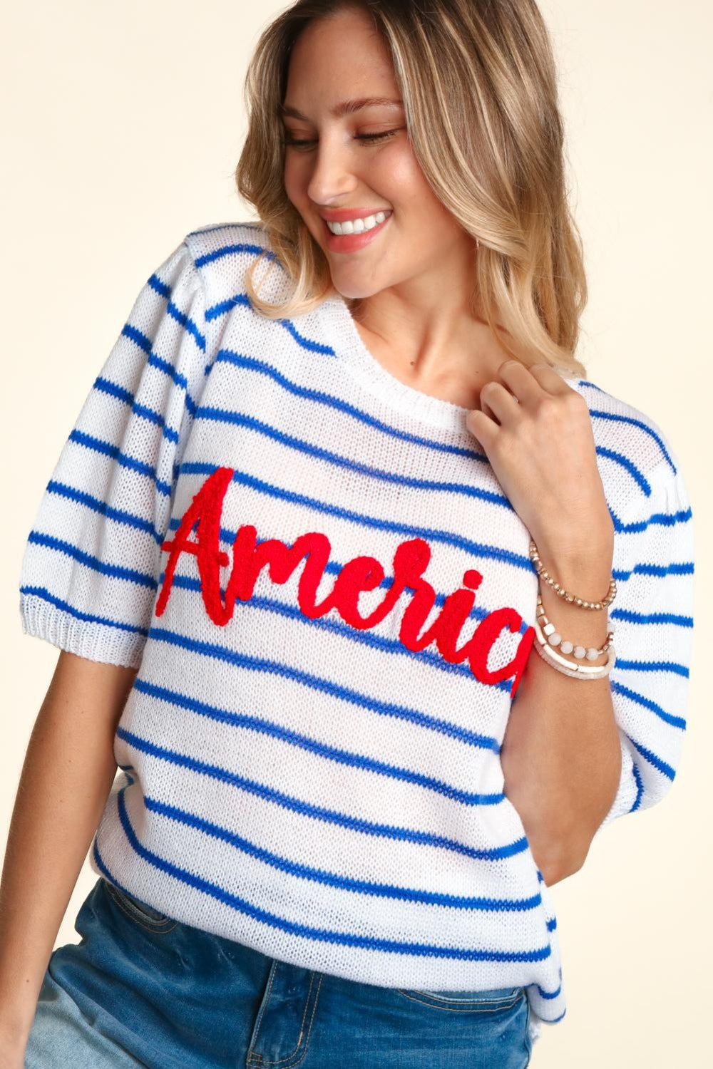 A woman wearing a chic Haptics Letter Embroidery Striped Knit Top featuring "America" in red stands against a neutral background. The white and blue striped knit design pairs perfectly with her denim shorts.