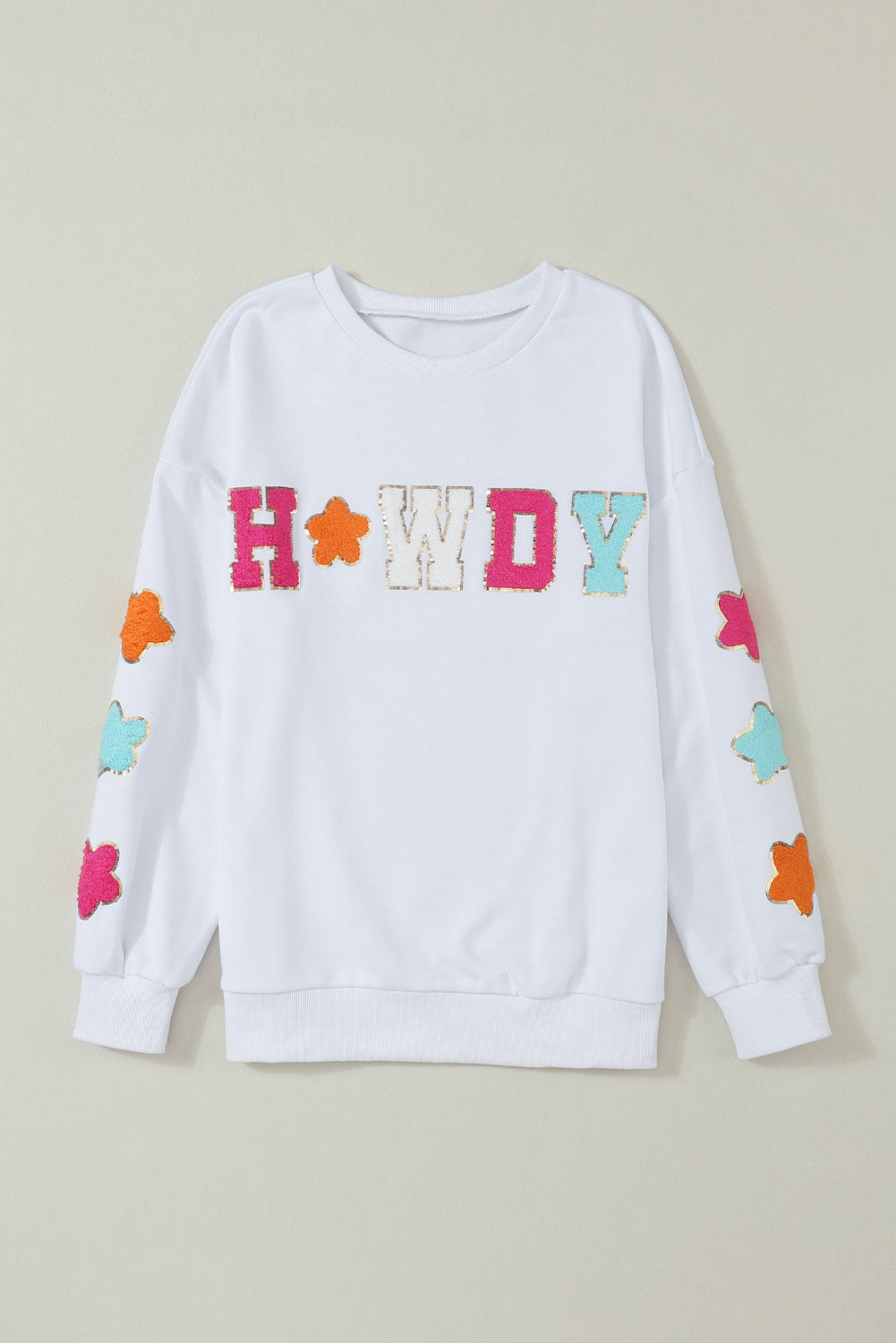 A person poses in the White Howdy Glitter Chenille Patch Graphic Casual Sweatshirt, adorned with colorful star patterns and the letters "WD." She wears light denim shorts and has shoulder-length blonde hair.