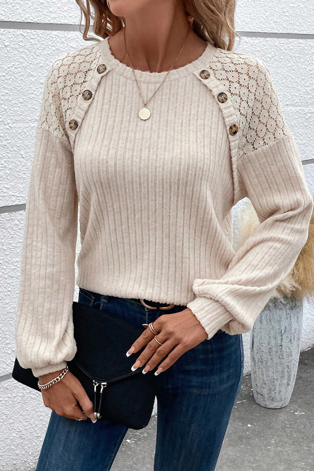 A woman dressed in the Parchment Contrast Lace Raglan Sleeve Buttoned Ribbed Top paired with blue jeans, a pendant necklace, and bracelets. She is holding a black clutch bag and has a hand on her waist.