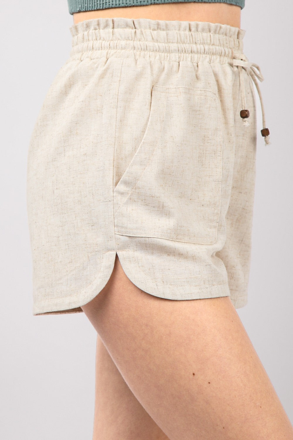 A person wearing VERY J Drawstring Elastic Waist Linen Shorts in beige and a green top. The image focuses on the midsection, with the eco-friendly shorts featuring front pockets and a slight texture.