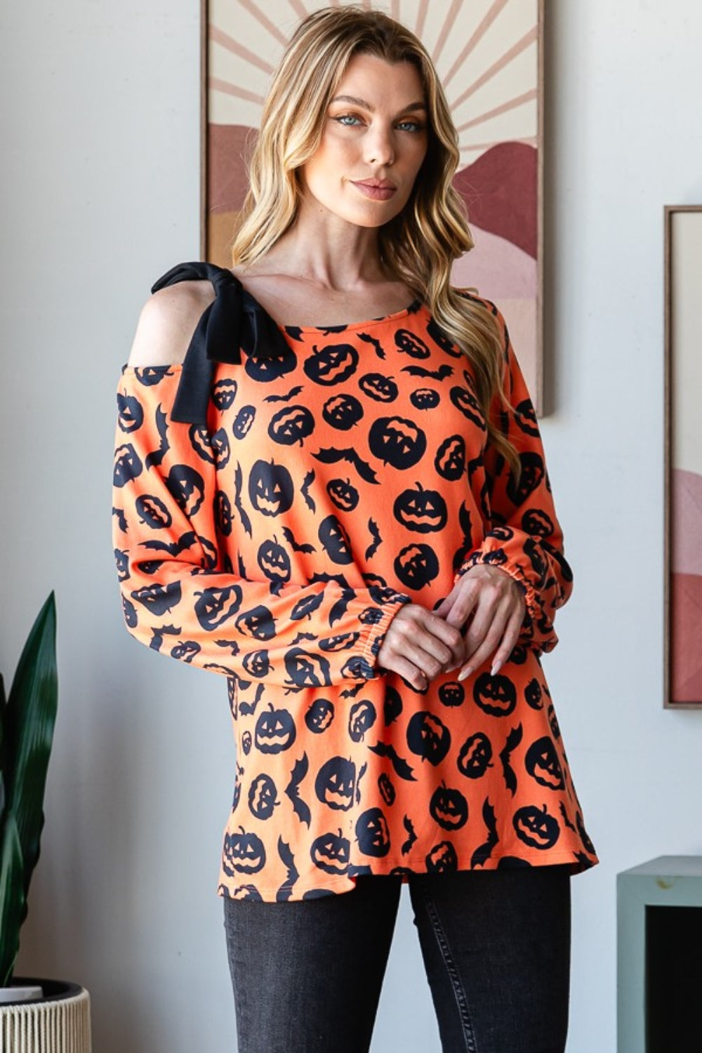 A woman smiles while modeling the Heimish Pumpkin Print One Shoulder T-Shirt, featuring an off-shoulder, long-sleeve design that is perfect for fall season fashion. She stands indoors with a plant and wall art in the background.