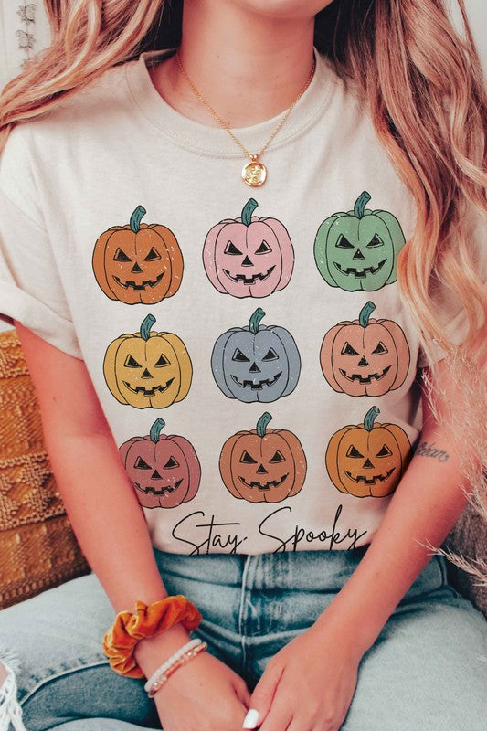 A person wears the STAY SPOOKY MULTI PUMPKINS Graphic Tee in unisex sizing, showcasing nine colorful jack-o'-lanterns and "Stay Spooky" text in a white hue, while holding a drink. They also sport checkered shorts.