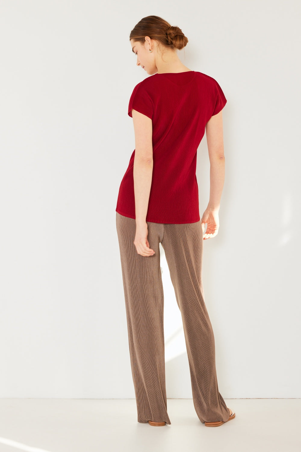 A person stands against a white wall, wearing a red short-sleeve shirt, Marina West Swim rib pleated elastic-waist wide-leg pants in beige, and white sandals. Their hands are in their pockets, and they sport a neutral expression.