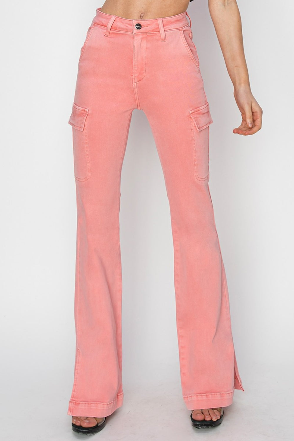 A person wearing RISEN Full Size High Rise Side Slit Cargo Bootcut Jeans, featuring a pink flared design with front pockets, stands against a plain background.