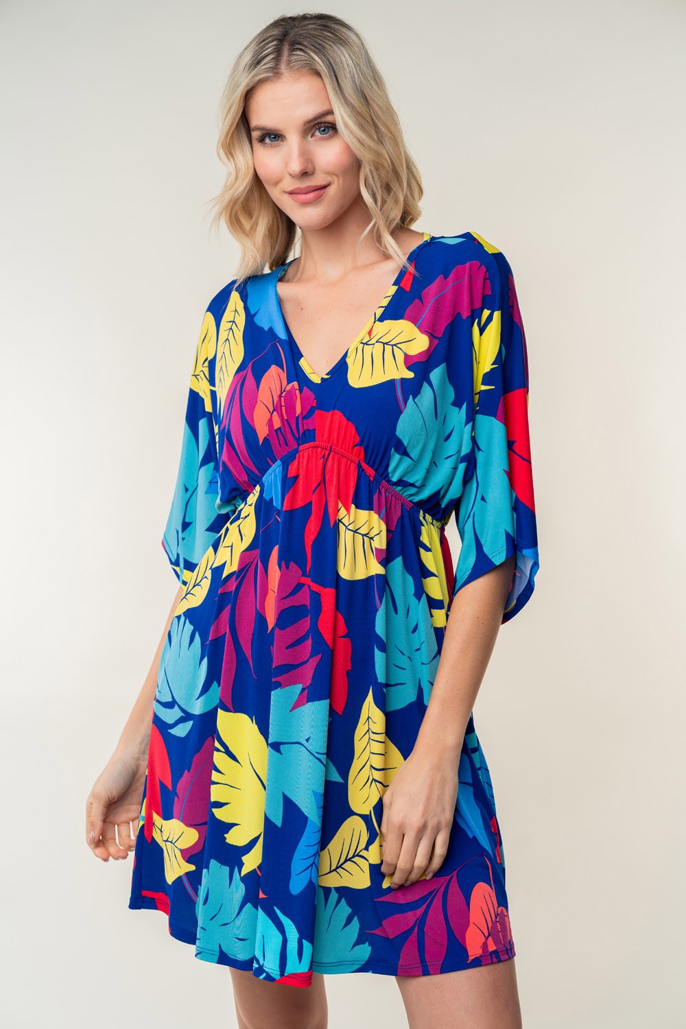 A person wearing the trendy White Birch Full Size Printed V-Neck Mini Dress, adorned with vibrant red, yellow, and blue leaves in a floral pattern, stands against a plain background.