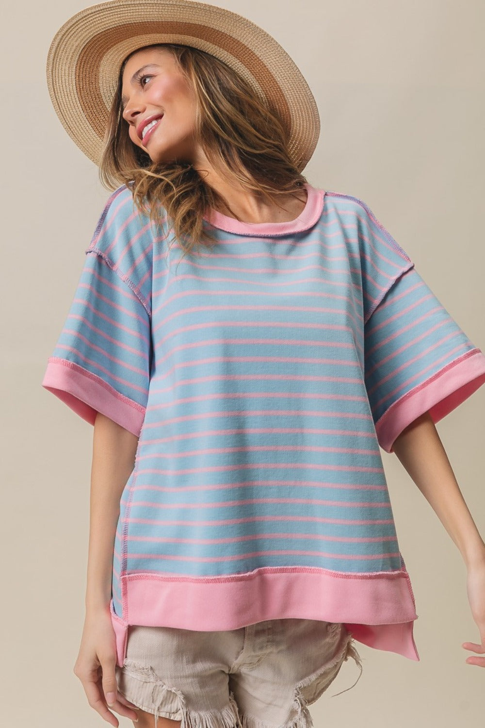 A person wearing the BiBi Exposed Seam Stripe Contrast T-Shirt in light blue and pink, paired with beige shorts and a wide-brimmed straw hat, stands against a plain background.