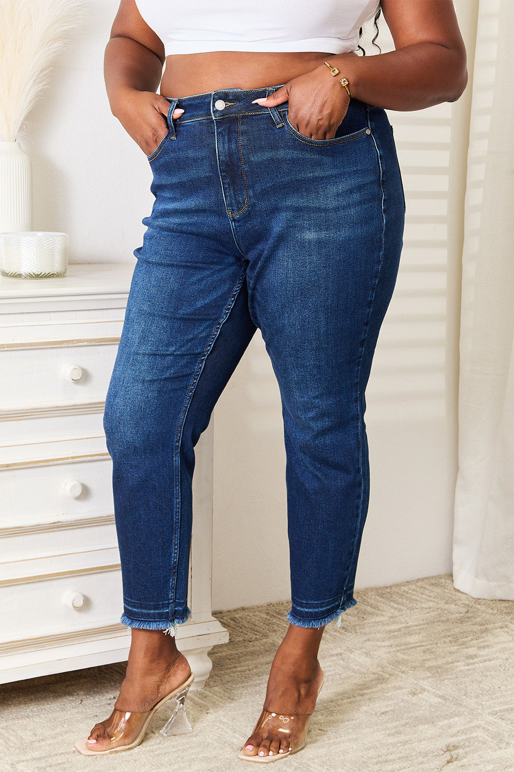 A person wearing Judy Blue Full Size High Waist Released Hem Slit Jeans in indigo wash and a white cropped top stands by a white dresser, showcasing a flattering fit.