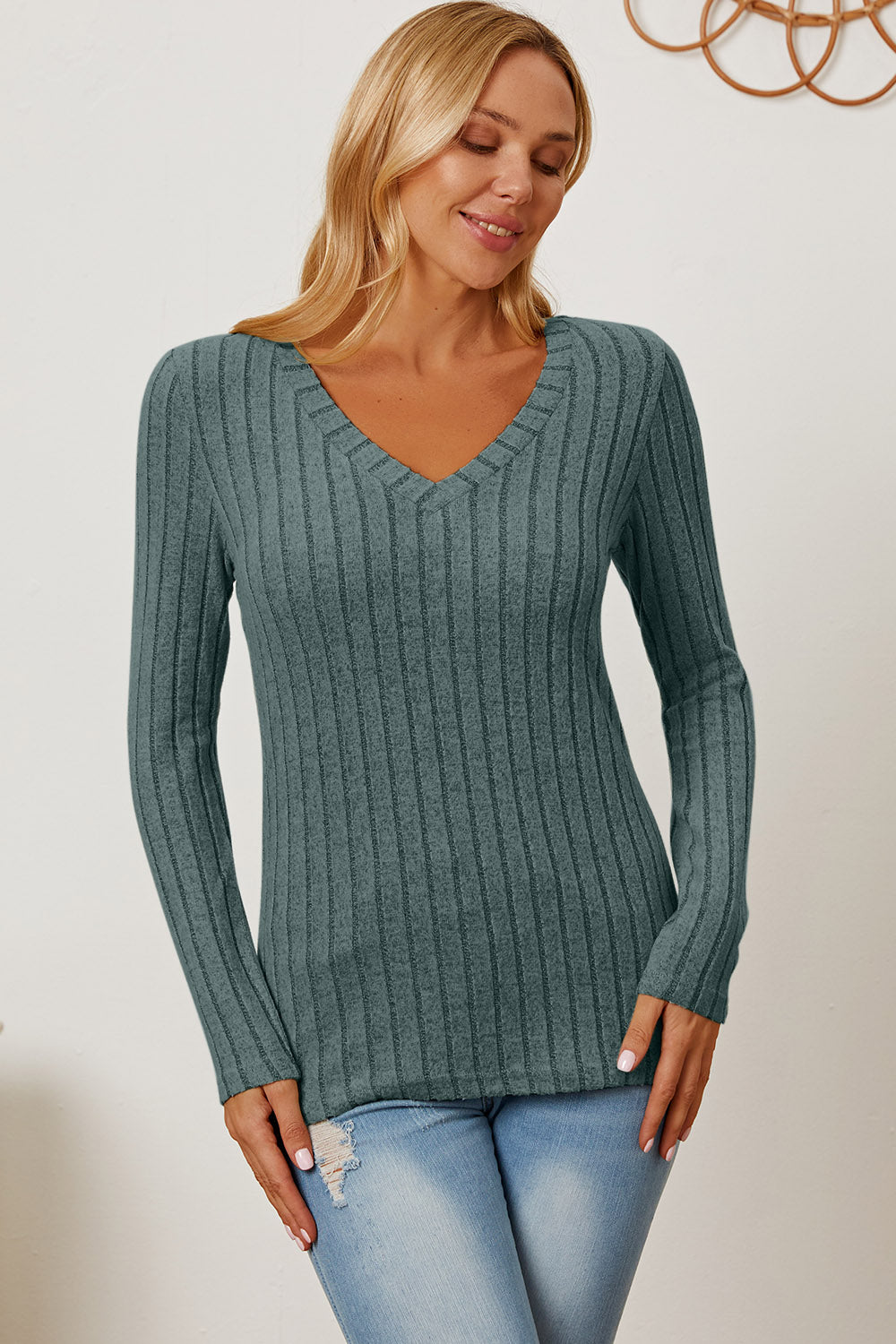 A blonde-haired woman smiles while wearing the Basic Bae Full Size Ribbed V-Neck Long Sleeve T-Shirt, which is light green and slightly stretchy, paired with light blue jeans. She holds one hand near her head and the other near her waist.