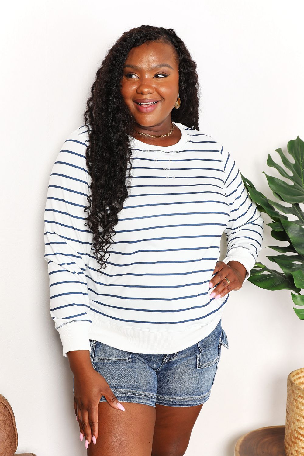 A person smiling while wearing the Double Take Striped Long Sleeve Round Neck Top, paired with denim shorts, stands in front of a white background with a plant.