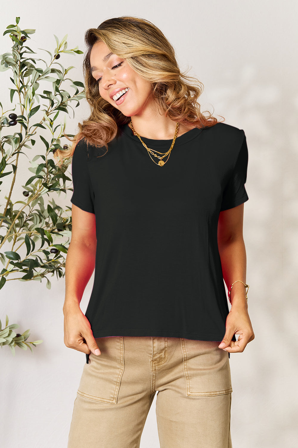 A woman with long hair, smiling with her right hand in her pocket and standing next to a plant, is wearing the Basic Bae Full Size Round Neck Short Sleeve T-Shirt in black along with beige pants made of rayon spandex.