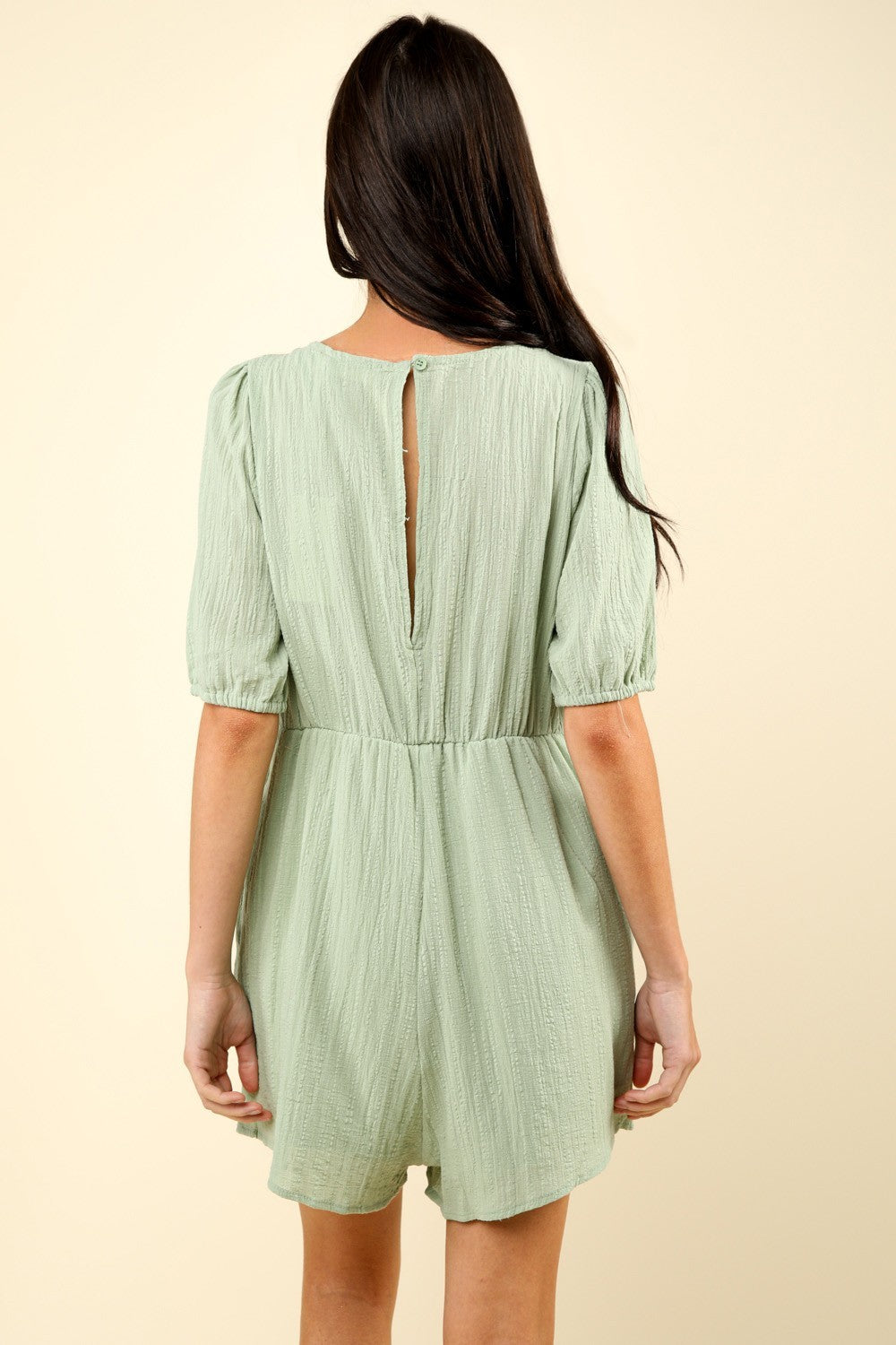 A person with long dark hair wearing a VERY J Lace Detail Puff Sleeve Romper with Pockets in light green, featuring a patterned bodice, stands against a plain beige background.