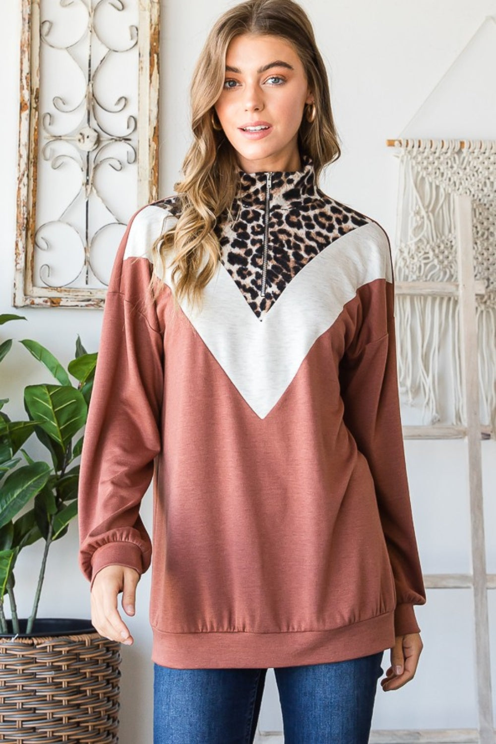 A woman wearing the stylish Heimish Full Size Leopard Quarter Zip Mock Neck Sweatshirt stands indoors near a lush plant and an intricate macrame wall hanging.