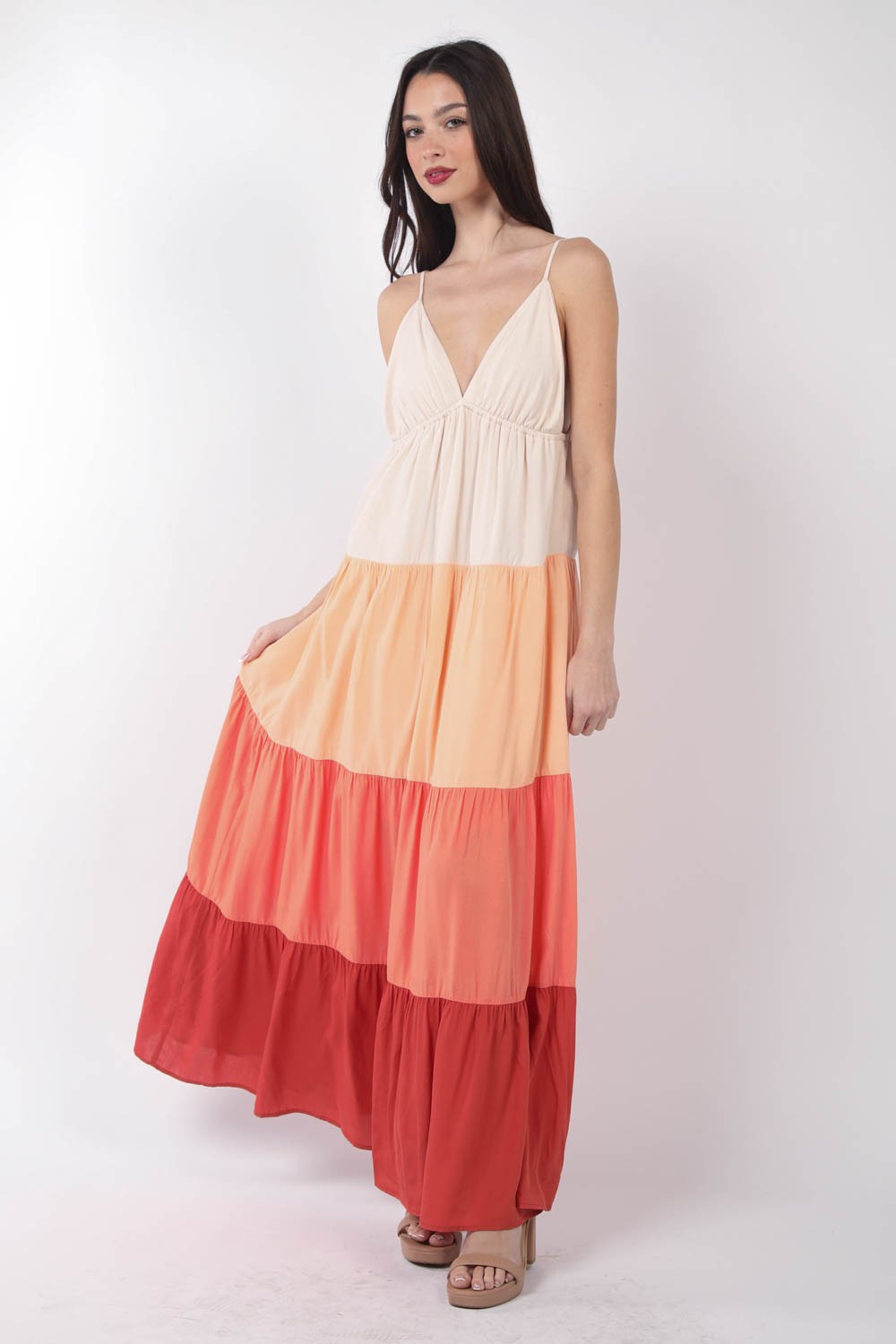 A woman stands, wearing the VERY J Color Block Tiered Maxi Cami Dress in shades of beige, peach, and red, paired with beige sandals. Her right hand holds the side of her dress as she poses against a plain white background—a truly fashionable choice.