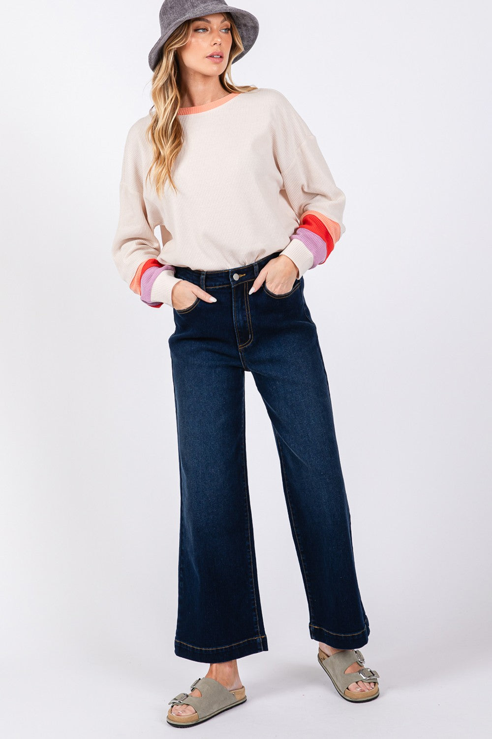 A person wearing a beige sweater, SAGE + FIG High Waist Wide Leg Jeans, a gray hat, and sandals is standing against a plain background.