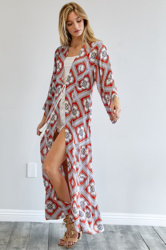 A person wearing a Printed Long Sleeve Loose Kimono with a bohemian blue and pink print over a white outfit stands in a room with a gray floor and white wall.