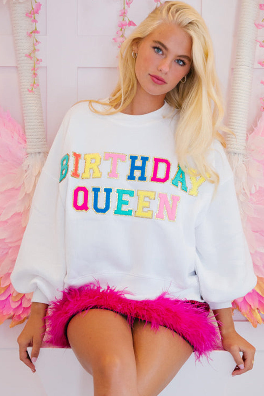 A person with long blonde hair is wearing a White BIRTHDAY QUEEN Graphic Balloon Sleeve Sweatshirt, paired with a pink feathered skirt, and sitting against a beautifully decorated backdrop.