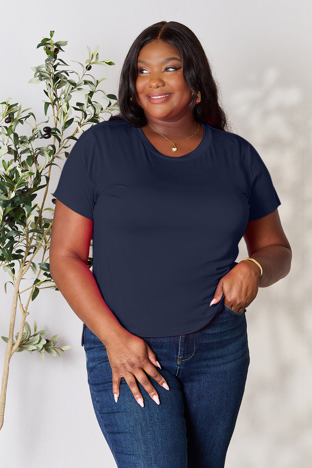 A person with wavy hair, wearing the Basic Bae Full Size Round Neck Short Sleeve T-Shirt in bright pink and beige pants, stands near a plant, smiling and looking to the side. The outfit embodies a basic style and offers slightly stretchy comfort, ideal for those who value effortless elegance.