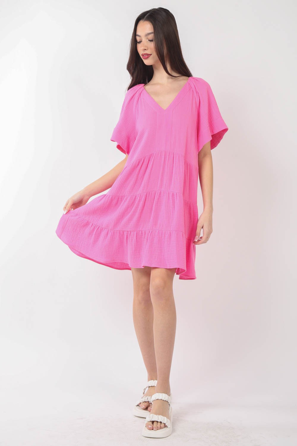 A woman is posing against a plain background, wearing the VERY J Texture V-Neck Ruffled Tiered Dress in pink, featuring short sleeves.