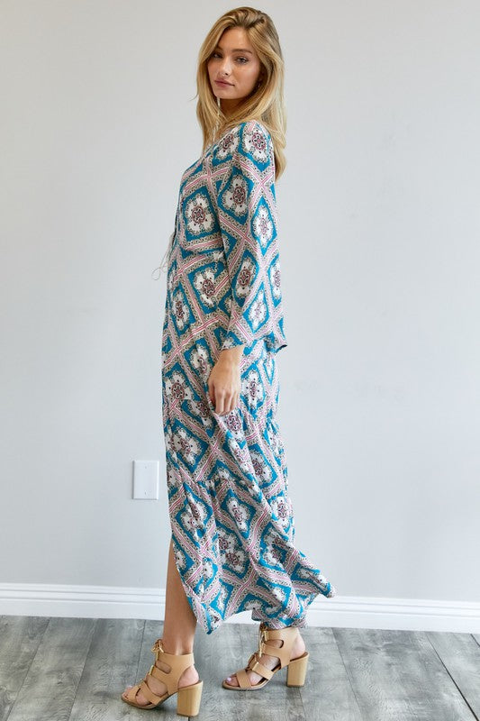 A person wearing a Printed Long Sleeve Loose Kimono with a bohemian blue and pink print over a white outfit stands in a room with a gray floor and white wall.