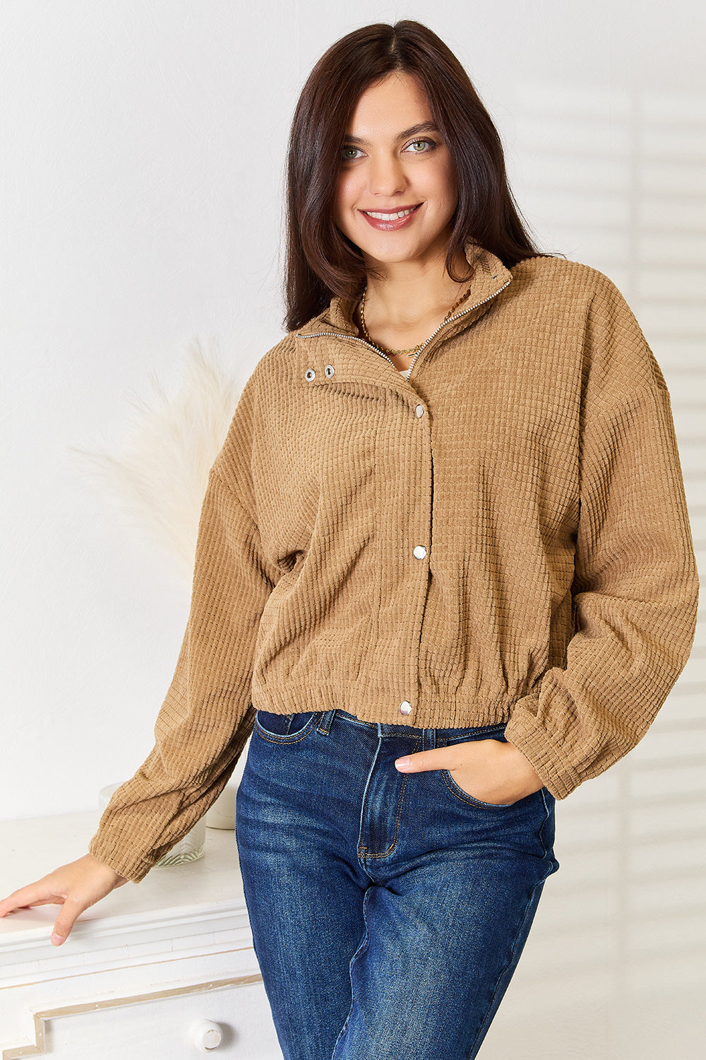 A person wearing the versatile Perfee Long Sleeve Dropped Shoulder Jacket in brown corduroy and blue jeans is standing indoors, smiling, with one hand resting on a white piece of furniture.