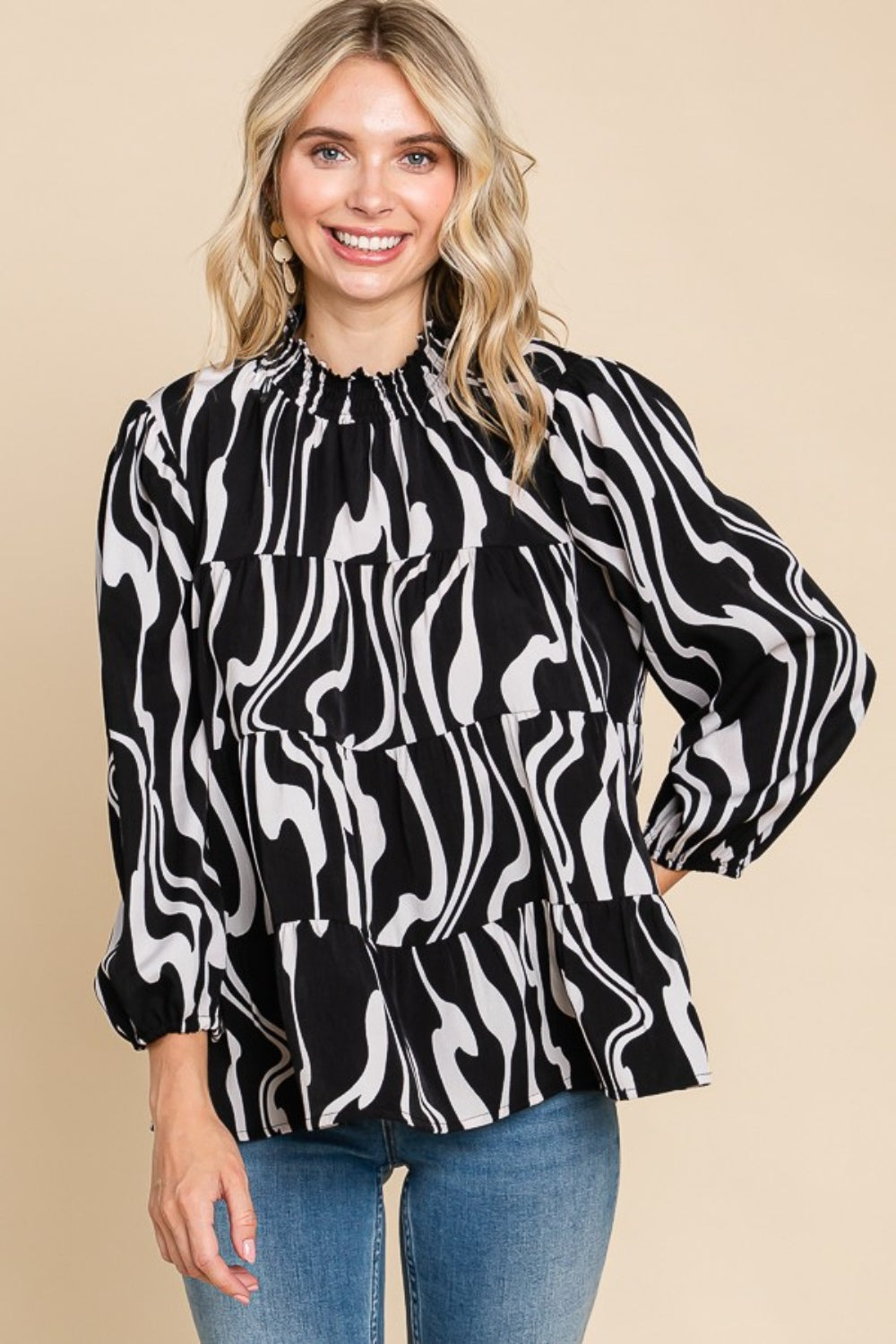 A person wearing a Culture Code Printed Smock Neck Tiered Blouse, featuring a black and white patterned design and long sleeves, paired with blue jeans, stands against a beige background.