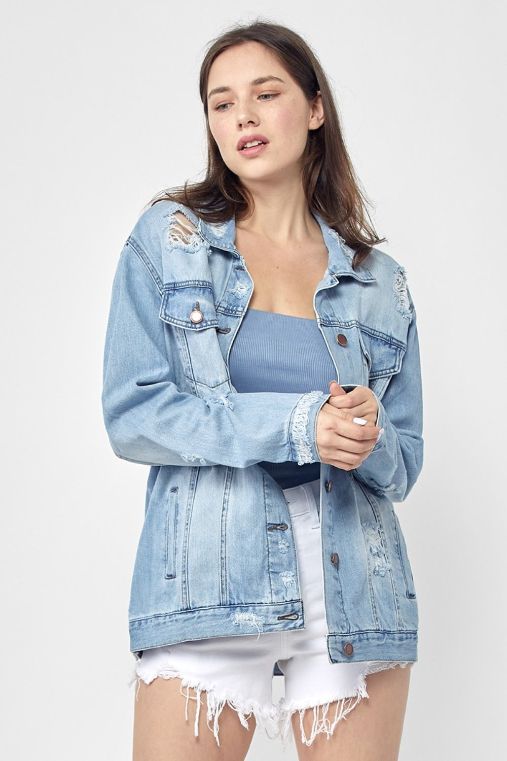 Dressed in the RISEN Full Size Distressed Long Sleeve Denim Jacket, a perfect wardrobe staple, this person effortlessly poses against a plain background while pairing it with a blue top and white shorts.