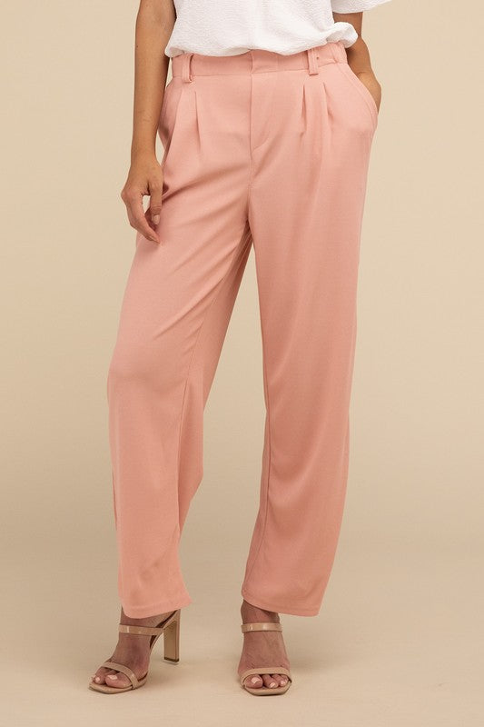 A woman stands against a beige background, highlighting her chic ensemble with a tailored fit. She wears a white short-sleeve shirt paired with versatile pink Waffle Trouser Pants, hands casually in pockets, complemented by stylish strappy sandals.